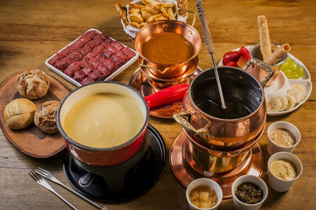 Panelas de fondue de queijo, carne e chocolate servidas em mesa de madeira